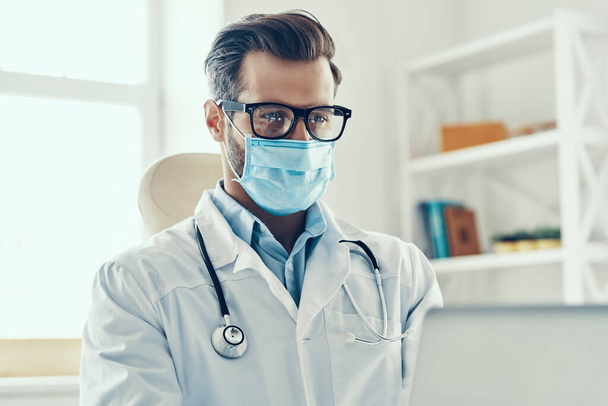 Dobře vypadající mladý muž v bílém laboratorním plášti a ochranné masky pracuje pomocí notebooku při sezení uvnitř - Fotografie, Obrázek