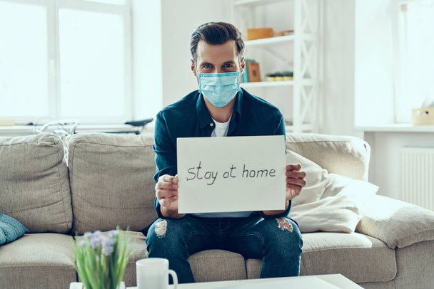 Young man in protective face mask showing stay at home saying written on the paper while sitting on sofa - Фото, зображення