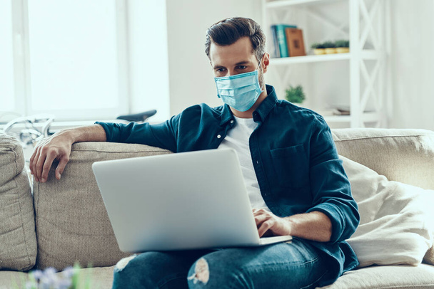 Concentrated young man in protective face mask using laptop while working from home - Photo, Image