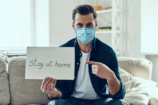 Handsome young man in protective face mask showing stay at home saying written on the paper while sitting on sofa - Foto, Bild
