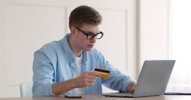 Młody człowiek wpisuje dane karty kredytowej i hasło bezpieczeństwa z telefonu komórkowego na laptopie - Materiał filmowy, wideo