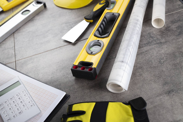 Contractor concept. Tool kit of the contractor: yellow hardhat, libella, hand saw. Plans and notebook on the gray tiles background. - Photo, Image