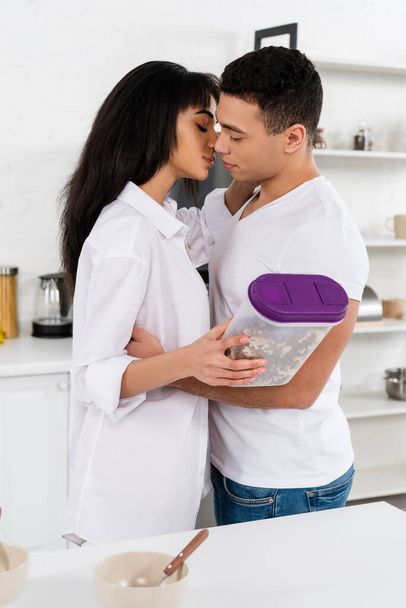 African american woman holding container with breakfast cereal, hugging and kissing with man near table in kitchen - Fotografie, Obrázek