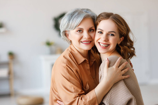 Cheerful young woman hugging aged mother sitting on sofa   while resting together in living room at hom - 写真・画像