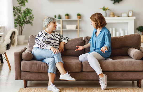 Полноразмерная молодая женщина улыбается и разговаривает со старшей матерью, сидя на удобном диване дома вместе с
 - Фото, изображение