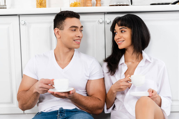 Interracial couple with saucers and cups of coffee smiling and looking at each other near kitchen cabinets - Photo, Image