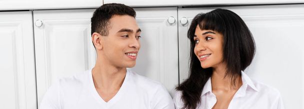 Межрасовая пара улыбается и смотрит друг на друга возле кухонных шкафов, панорамный снимок
 - Фото, изображение
