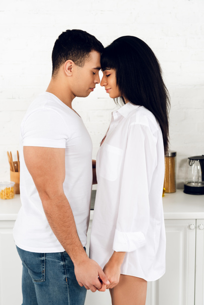 Interracial couple with closed eyes facing each other and holding hands in kitchen - Photo, image