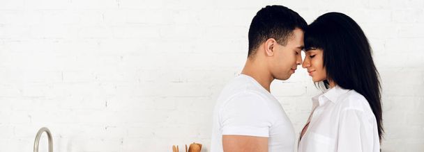 Casal inter-racial com os olhos fechados de frente para o outro e sorrindo na cozinha, tiro panorâmico
 - Foto, Imagem