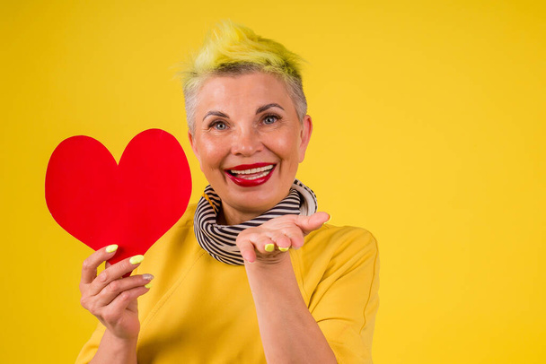 mature old woman with colored stylish yellow dyed hair and red lipstick lips in warm dress holding paper Valentine card background studio - Fotoğraf, Görsel