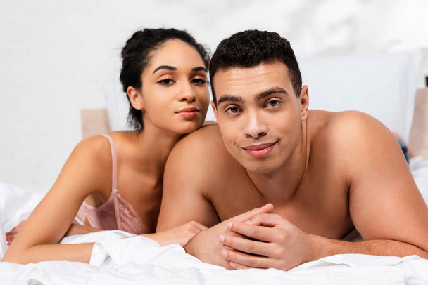Mężczyzna z zaciśniętymi rękami w pobliżu afrykańskiej amerykańskiej kobiety patrzącej w kamerę i leżącej na łóżku w sypialni - Zdjęcie, obraz