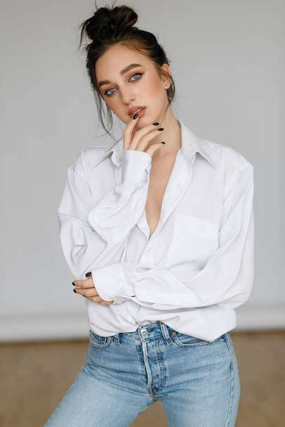 portrait of seated young woman wearing a bouffant shirt with naked shoulders on light grey background. Beautiful brunette girl in jeans and white t-shirt smiling - Фото, зображення