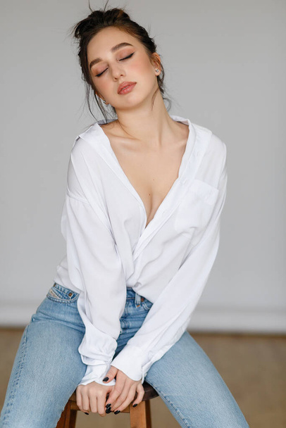 portrait of seated young woman wearing a bouffant shirt with naked shoulders on light grey background. Beautiful brunette girl in jeans and white t-shirt smiling - Fotografie, Obrázek
