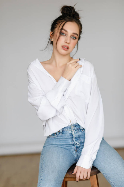 portrait of seated young woman wearing a bouffant shirt with naked shoulders on light grey background. Beautiful brunette girl in jeans and white t-shirt smiling - Fotoğraf, Görsel