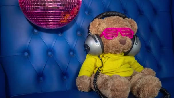 пушистая игрушка медвежонка в солнцезащитных очках и наушниках с диско шаром на диване
 - Кадры, видео