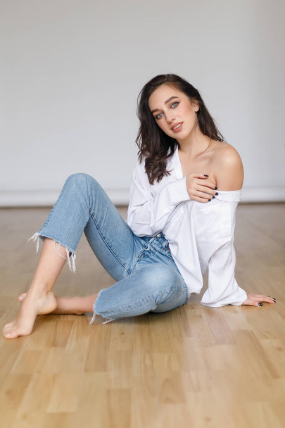 portrait of seated young woman wearing a bouffant shirt with naked shoulders on light grey background. Beautiful brunette girl in jeans and white t-shirt smiling - Zdjęcie, obraz
