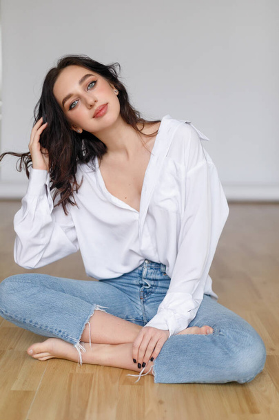 portrait of seated young woman wearing a bouffant shirt with naked shoulders on light grey background. Beautiful brunette girl in jeans and white t-shirt smiling - Foto, imagen