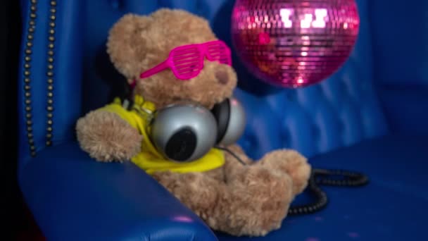 bruine teddybeer pluizig speeltje in zonnebril en hoofdtelefoon met discobal op sofa achtergrond - Video