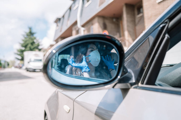 Pohled do zpětného zrcátka auta, ve kterém je vidět atraktivní žena v lékařských rukavicích, jak si nasazuje masku proti chřipce.Kopírovat prostor.Selektivní soustředění.Zdravotní koncepce, Covid-19 - Fotografie, Obrázek
