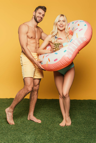 happy blonde girl in swim ring holding pineapple with cocktail near shirtless muscular man on yellow background - Photo, image