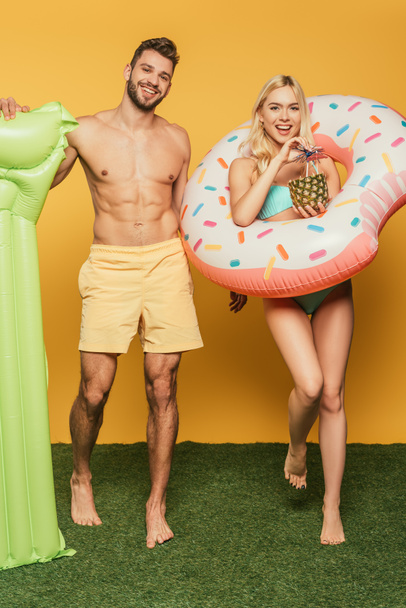 handsome muscular man with inflatable mattress and smiling blonde girl with swim ring and pineapple with cocktail on yellow background - Foto, immagini