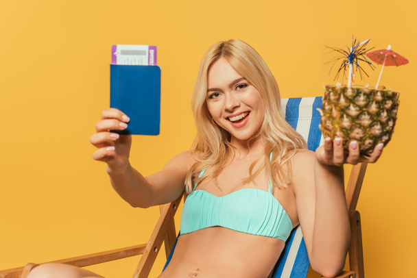 happy blonde girl in swimsuit holding documents and half of fresh pineapple while sitting in deck chair on yellow background - Foto, Imagem
