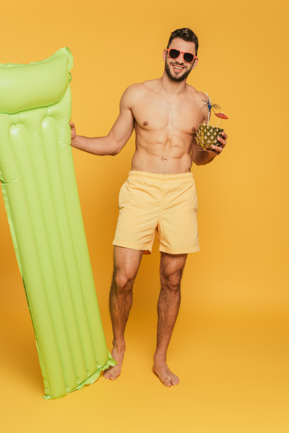 full length view of cheerful muscular man holding inflatable mattress and half of pineapple with fresh cocktail on yellow background - 写真・画像