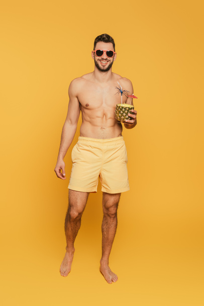 Ganzkörperansicht eines lächelnden, hemdslosen Mannes mit Sonnenbrille, der die Hälfte der Ananas mit frischem Cocktail auf gelbem Hintergrund hält - Foto, Bild