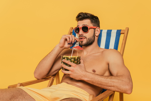 handsome shirtless man in sunglasses drinking fresh cocktail from half of pineapple while relaxing in deck chair isolated on yellow - 写真・画像