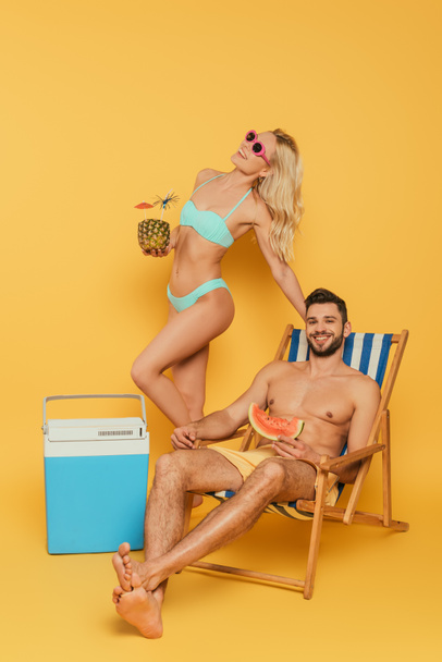 attractive blonde girl holding pineapple with cocktail near portable fridge, and smiling man sitting in deck chair on yellow background - Фото, зображення