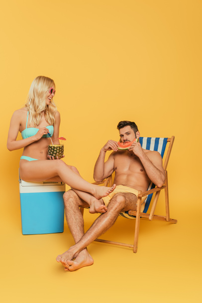 smiling blonde girl sitting on portable fridge near handsome man eating watermelon in deck chair on yellow background - Фото, изображение