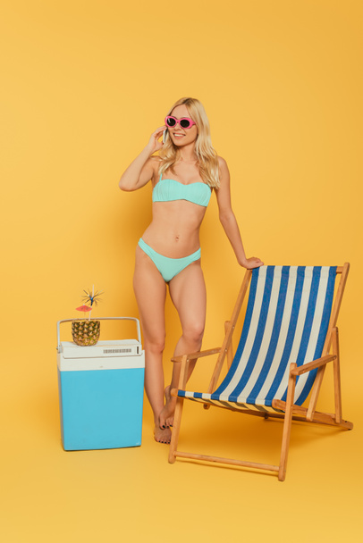 smiling, blonde girl talking on smartphone near deck chair and portable fridge with fresh cocktail in pineapple on yellow background - Fotó, kép