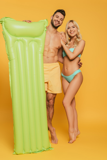 happy muscular man holding inflatable mattress and hugging attractive blonde girl on yellow background - Foto, immagini