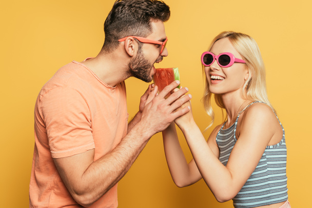 cheerful blonde girl feeding handsome boyfriend with juicy watermelon on yellow background - Valokuva, kuva