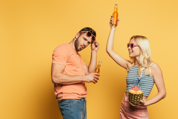 aufgeregtes junges Paar mit Sonnenbrille und Cocktail in Ananas und Bierflaschen auf gelbem Hintergrund - Foto, Bild