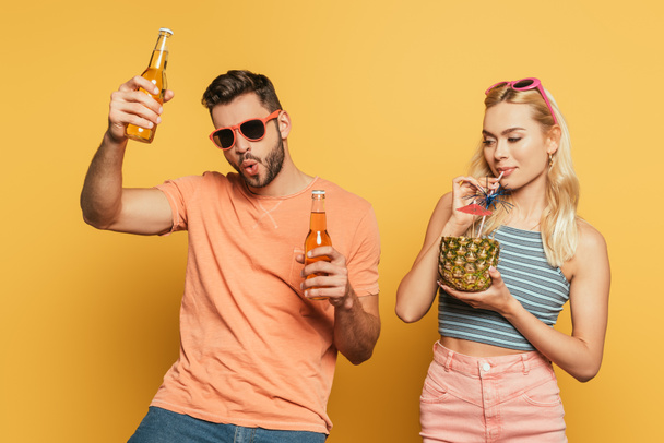 homme excité avec des bouteilles de bière près de fille blonde boire cocktail de l'ananas sur fond jaune
 - Photo, image