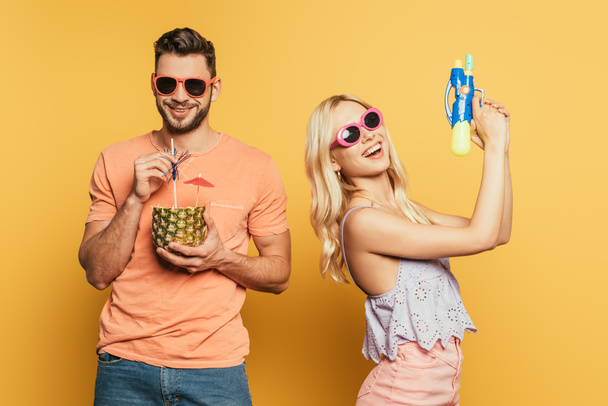 cheerful blonde girl holding water gun near smiling man drinking cocktail from half of pineapple on yellow background - Fotó, kép
