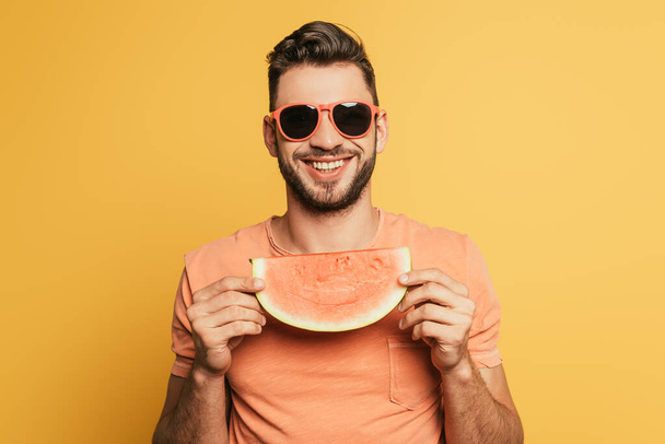 щасливий молодий чоловік в сонцезахисних окулярах тримає шматочок стиглого кавуна на жовтому фоні
 - Фото, зображення