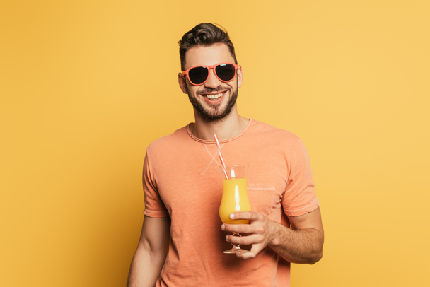 fröhlicher junger Mann mit Sonnenbrille lächelt in die Kamera, während er ein Glas Orangensaft auf gelbem Hintergrund hält - Foto, Bild