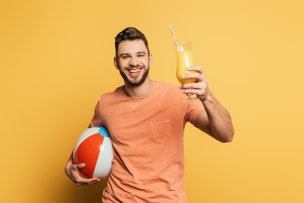 happy young man holding inflatabe ball and glass of orange juice on yellow background - Foto, afbeelding