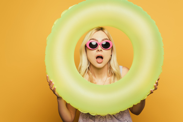 shocked blonde girl with open mouth looking through swim ring on yellow background - Photo, Image