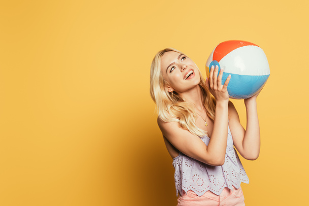 happy, blonde girl holding inflatable ball while looking up on yellow background - Valokuva, kuva