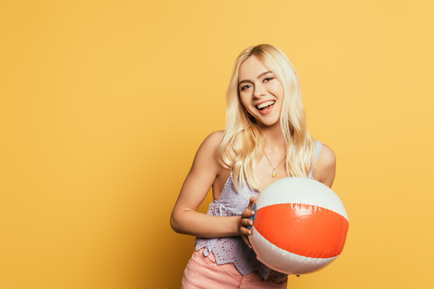 attractive, blonde girl holding inflatable ball while smiling at camera on yellow background - Fotografie, Obrázek