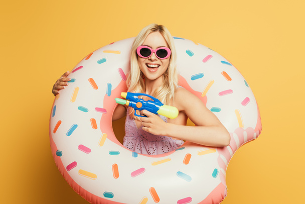 smiling blonde girl in swim ring holding water gun on yellow background - 写真・画像