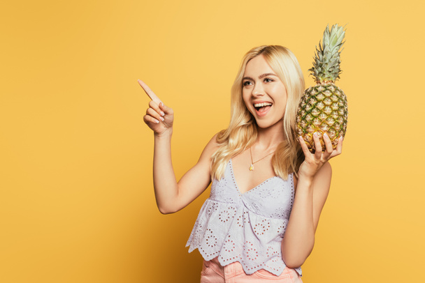 cheerful blonde girl holding pineapple and pointing with finger on yellow background - Foto, afbeelding