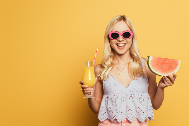 happy blonde girl in sunglasses holding orange juice and slice of watermelon on yellow background - Fotó, kép