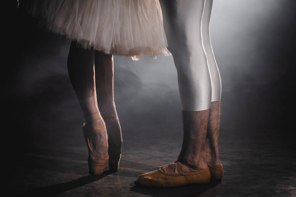 Close up of ballet couple of dancers as they practices exercises on dark stage or studio. Womans and mans feet in pointe shoes. Ballerina shows classic ballet pas. Slow motion. Flare shot. - Foto, imagen