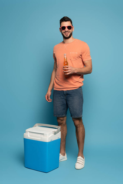 happy man in sunglasses holding bottle of beer while standing near portable fridge on blue background - Photo, Image