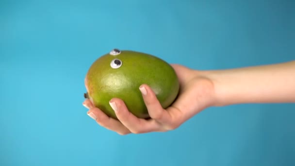 Mango apple with eyes in a woman hand close up. Apple looks around on a blue background. Vertical video. - Footage, Video