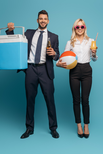 volledige weergave van zakenman met draagbare koelkast en bier in de buurt van lachende zakenvrouw met opblaasbare bal en sinaasappelsap op blauwe achtergrond - Foto, afbeelding